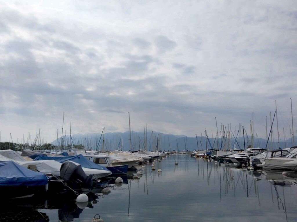 Boote und Segelmasten im Yachthafen von Ouchy mit Genfersee und Alpen im Hintergrund.