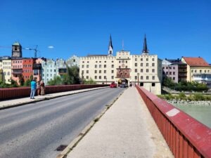 Blick über die rote Innbrücke auf das Brucktor und die pastellfarbenen Häuser der Wasserburger Altstadt bei blauem Himmel.