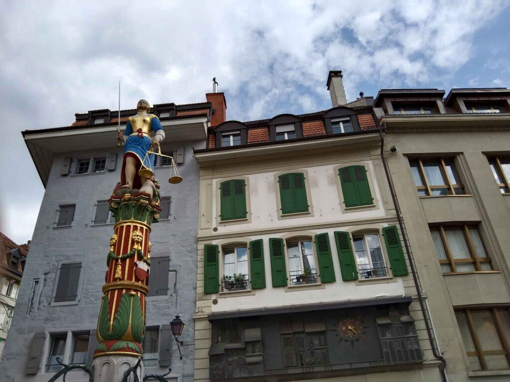 Farbig bemalte Justitiafigur mit Schwert und Waage auf dem Renaissancebrunnen am Place de la Palud vor historischen Häusern.