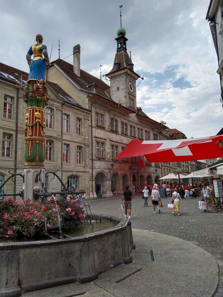 Place de la Palud in Lausanne mit Renaissancebrunnen, Justitiafigur, Rathaus und belebtem Platz unter rot-weißen Sonnensegeln.