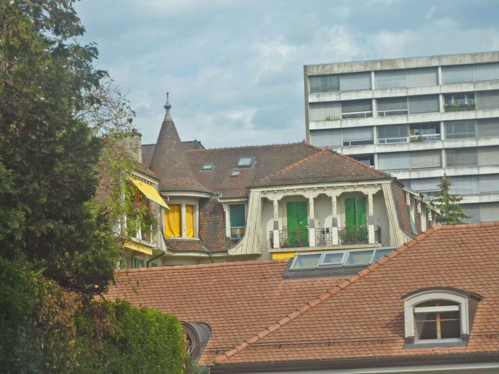 Verschachtelte Ziegeldächer mit Gauben, kleinem Turm, Balkonen und Fensterläden vor einem modernen Wohnhaus in Lausanne.