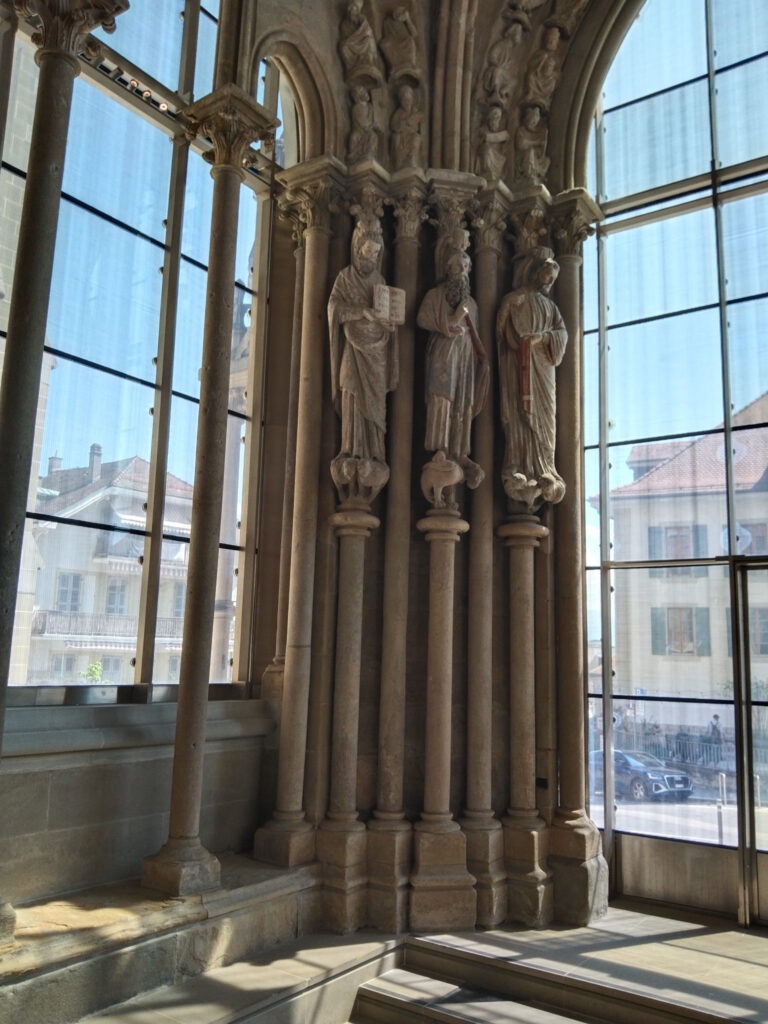Radsport, Hitze und Erbauung 11 Drei mittelalterliche Steinfiguren zwischen schlanken Säulen im Eingangsbereich der Kathedrale von Lausanne.