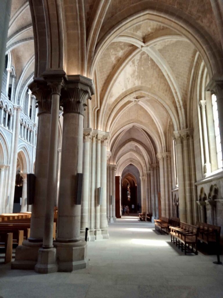 Langes Seitenschiff der Kathedrale von Lausanne mit Säulen, Spitzbögen, Kreuzrippengewölbe und Sitzbänken.