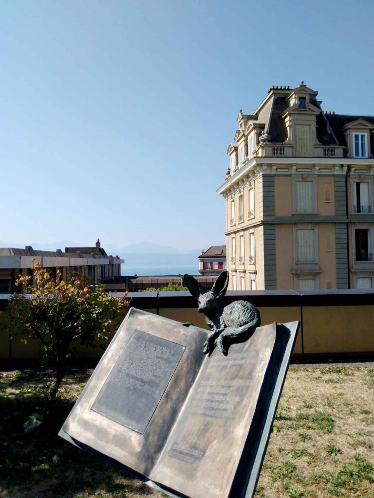 Bronzefarbene Fuchsskulptur auf einem aufgeschlagenen Buch im Park von Montbenon mit Gebäuden und Genfersee im Hintergrund.