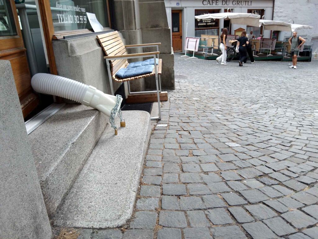 Weißer Abluftschlauch führt aus einer Ladentür auf einen gepflasterten Gehweg und ist mit einer Kette gesichert.