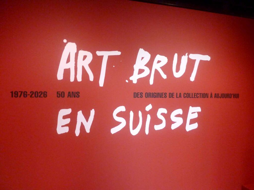 Rote Ausstellungswand mit der weißen Aufschrift Art Brut en Suisse und dem Hinweis 1976–2026, 50 ans.