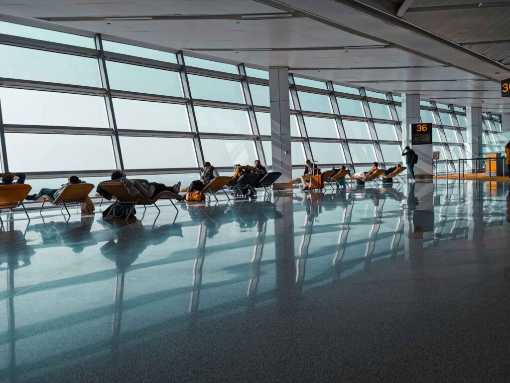 Schlafen am Flughafen: Wo Reisende in Europa am besten übernachten 6 Reisende ruhen auf Liegesesseln in einem hellen Flughafenterminal mit großer Fensterfront und spiegelndem Boden